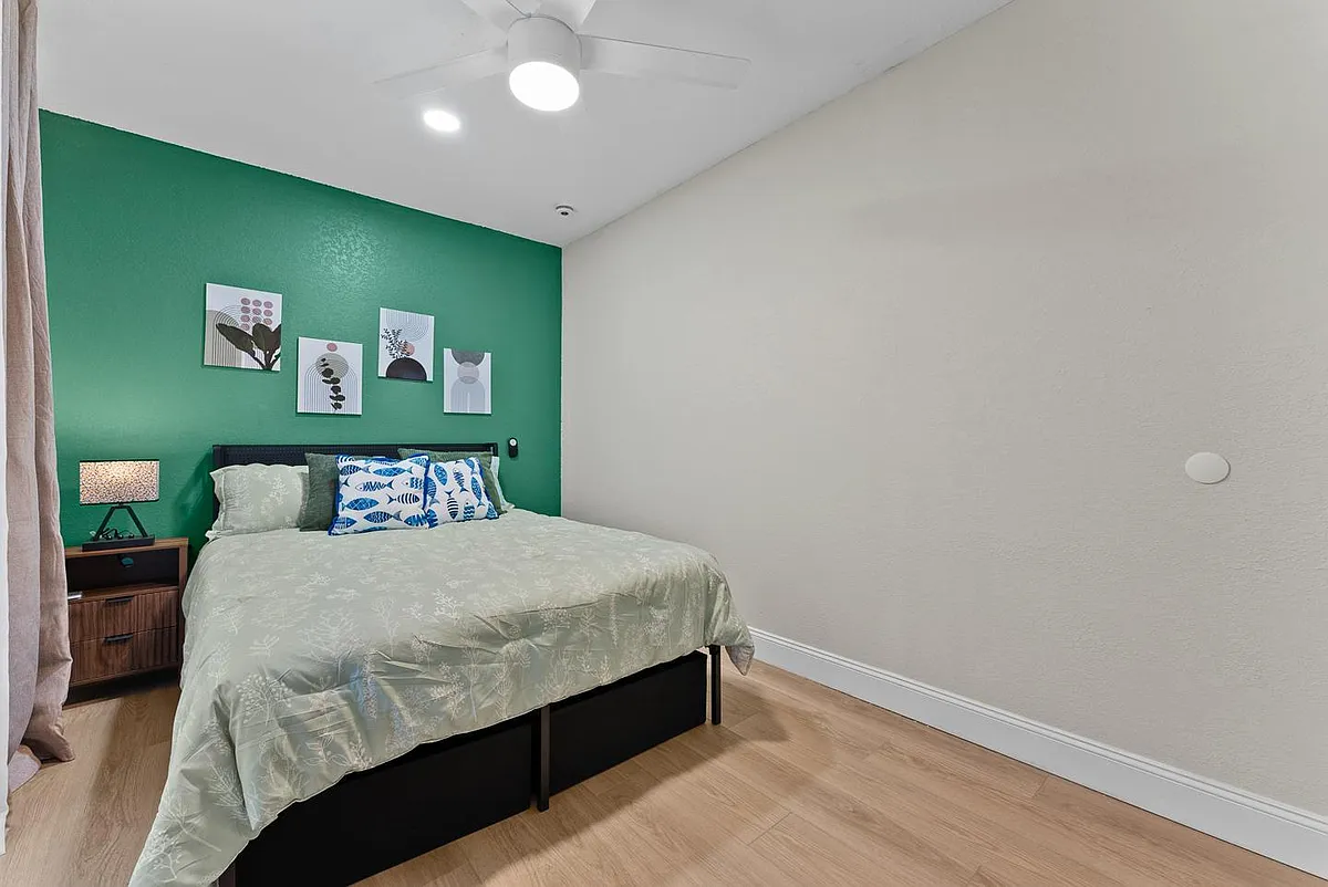 Furnished bedroom with a full-size bed, bedside table and lamp, ceiling fan, and window with curtains for privacy. - 12th Ave E