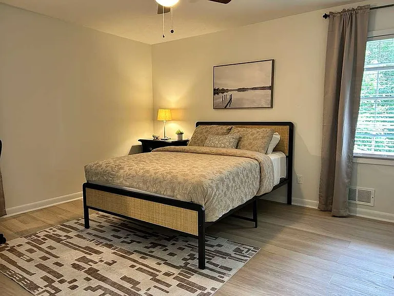 Furnished bedroom with a full bed, nightstand lighting, and a large window with blinds and curtains for privacy.