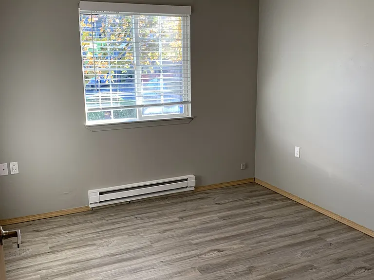 Bright bedroom with wood-style flooring, neutral tones, and a large window for natural light. Offers a clean, versatile space perfect for a bedroom, home office, or flex use.