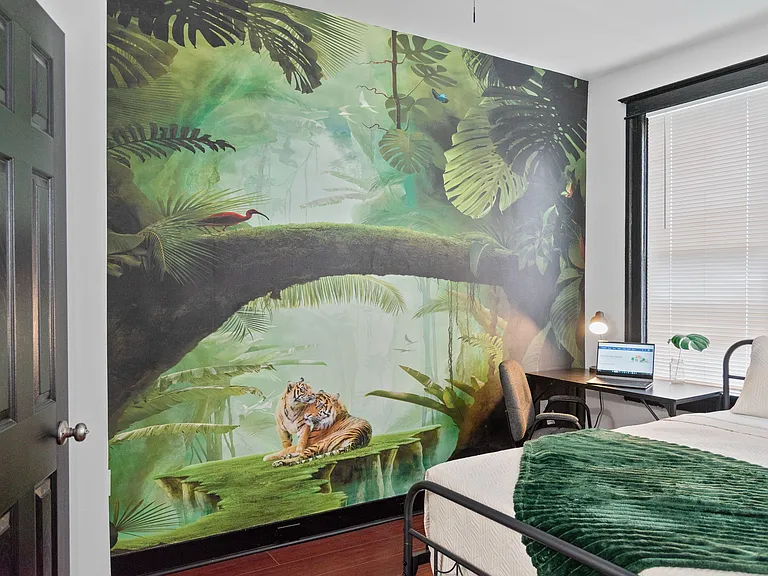 Furnished bedroom with twin bed, dedicated desk workspace, and large window with blinds plus a bold accent mural.