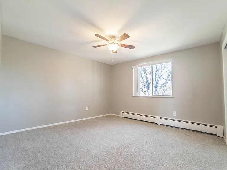Bright and spacious bedroom with plush carpeting and a ceiling fan, featuring a large window with a serene view.