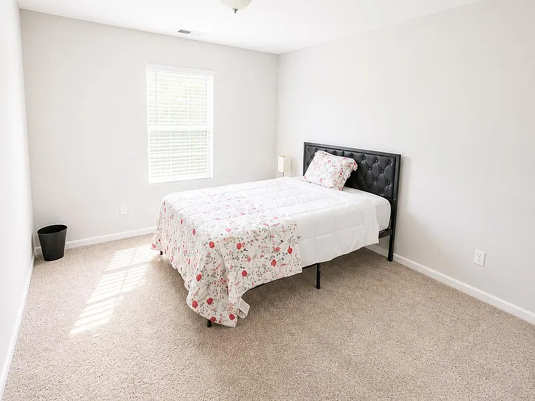Spacious carpeted bedroom with a large window, neutral walls, and room for additional furniture or a work desk.