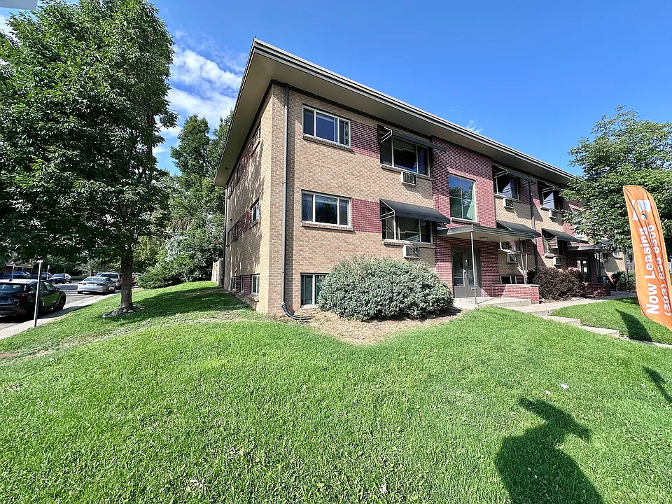 Charming two-story brick apartment building with lush green lawns, large windows, and a welcoming entrance. Now leasing sign signifies immediate availability in this appealing neighborhood.