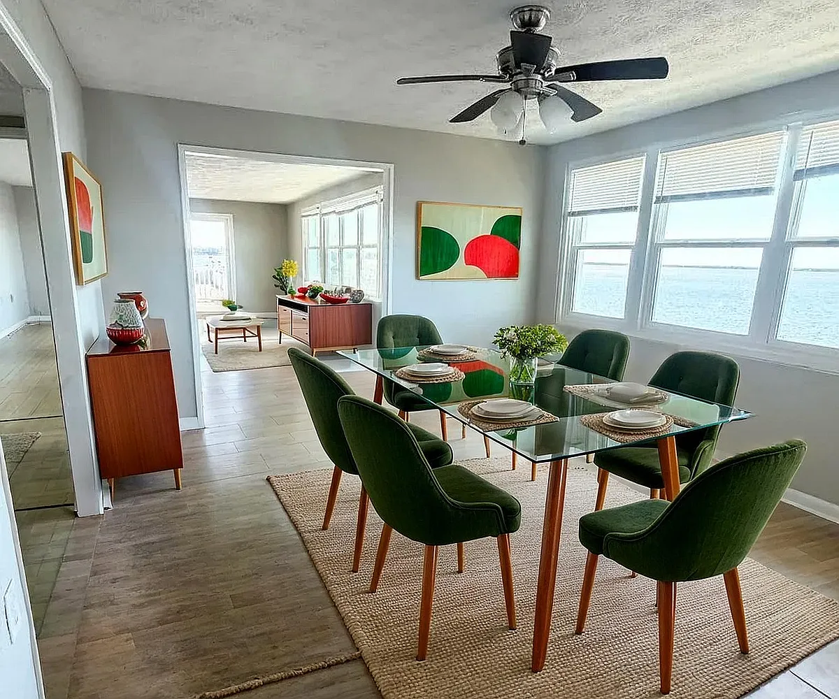 Chic dining area with green velvet chairs and glass table, showcasing modern decor and abundant natural light with stunning ocean views. - River Road Apartments