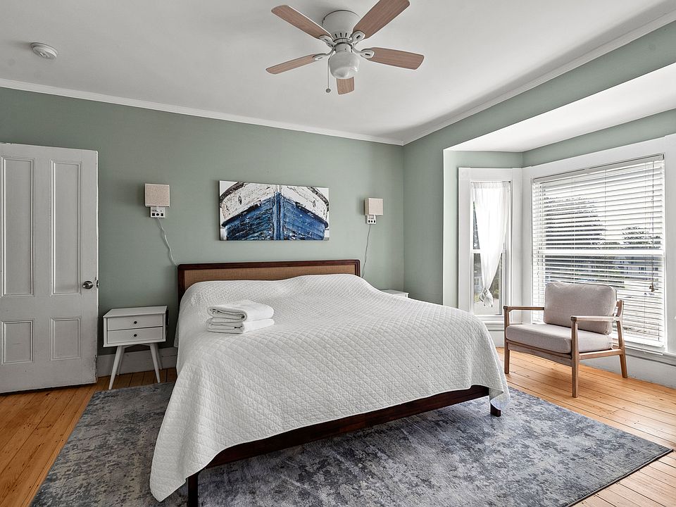 The focal point is a comfortable king-sized bed dressed in crisp white linens, centered beneath a unique nautical-themed art piece. The walls are painted in a soothing, calming sage green. Enjoy the sun-drenched bay window nook, featuring a cozy reading c