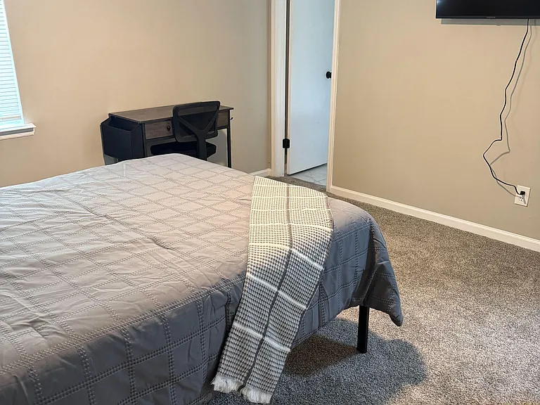 Furnished bedroom with a comfortable bed, wall-mounted TV, and dedicated desk area, plus carpeted floors and a window for lig