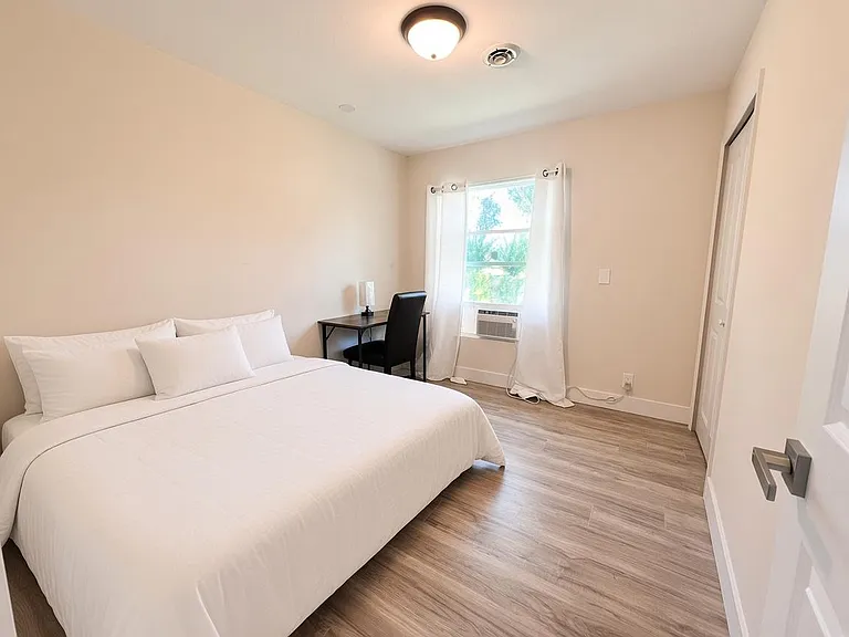 Spacious furnished bedroom with wood-look flooring, a work desk, and a window with AC for comfortable year-round living.