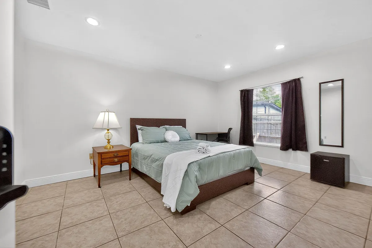 Spacious furnished bedroom with tile flooring, recessed lighting, a work desk, and a large window with curtain panels. - SW 43rd Terrace Rd