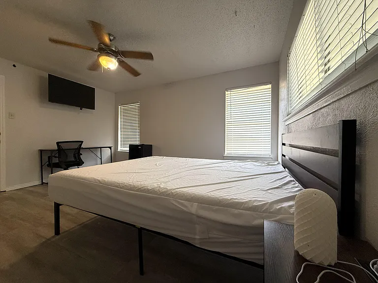 Furnished bedroom with bed frame, wall-mounted TV, dedicated desk area, and windows with blinds for natural light.