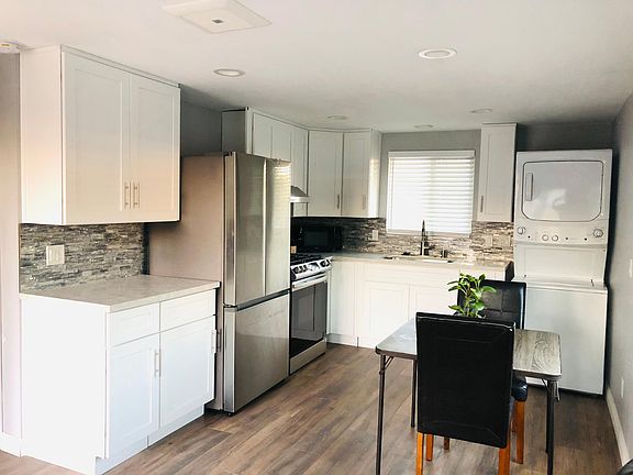 New white solid wood kitchen cabinets with quartz countertop and stainless steel appliances (refrigerator, kitchen hood, stove, stacked washer/dryer), dining table with 2 chairs