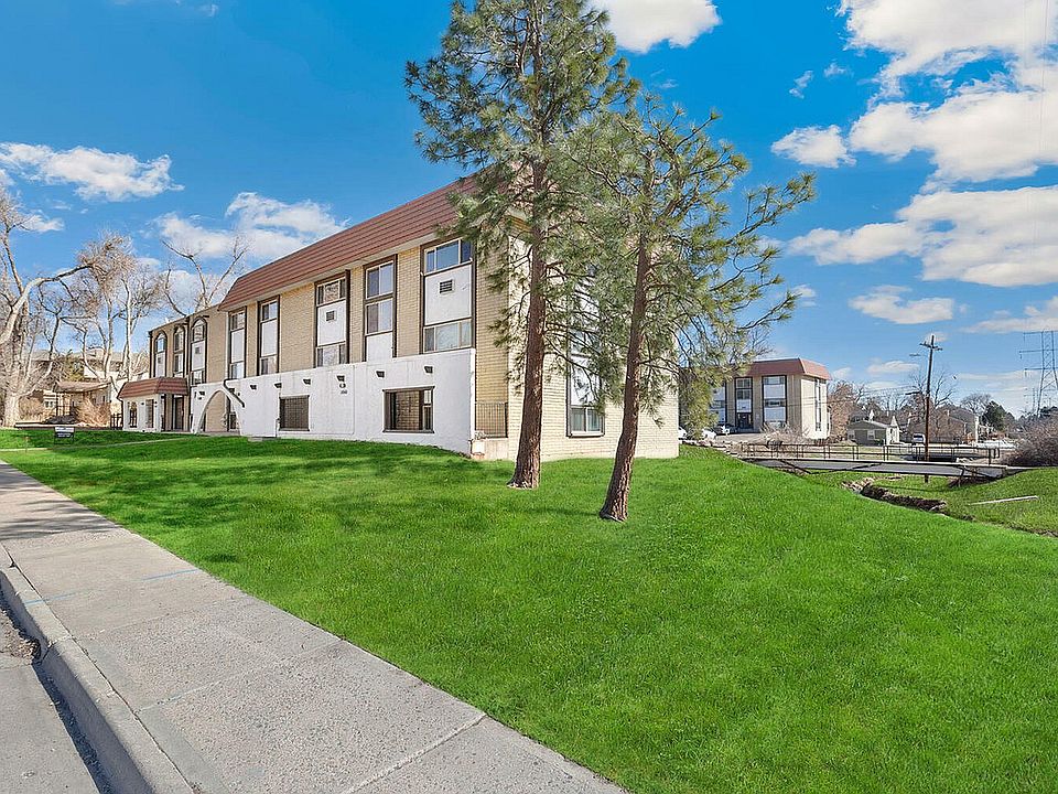 Charming apartment building surrounded by lush green lawns and picturesque trees under a vibrant blue sky.