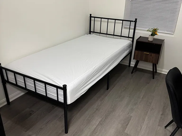 Furnished private bedroom with a twin bed, nightstand, and a window with blinds for light and privacy.