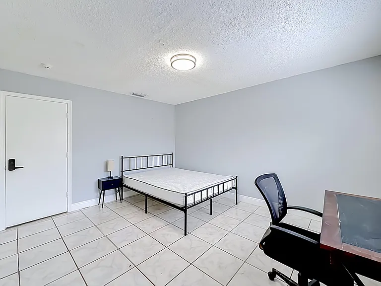 Spacious bedroom with tiled floors, a bed frame, nightstand, and a dedicated desk area for work or study.