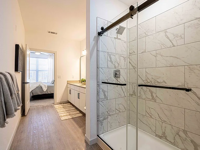 The primary bathroom shower in this Garrett Parker B1 floor plan includes floor to ceiling marble tile, a glass sliding door, and a rainfall shower head.