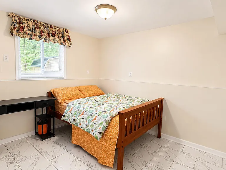 Furnished bedroom with a full bed, side workspace, and tile flooring, plus a window with blinds for natural light.