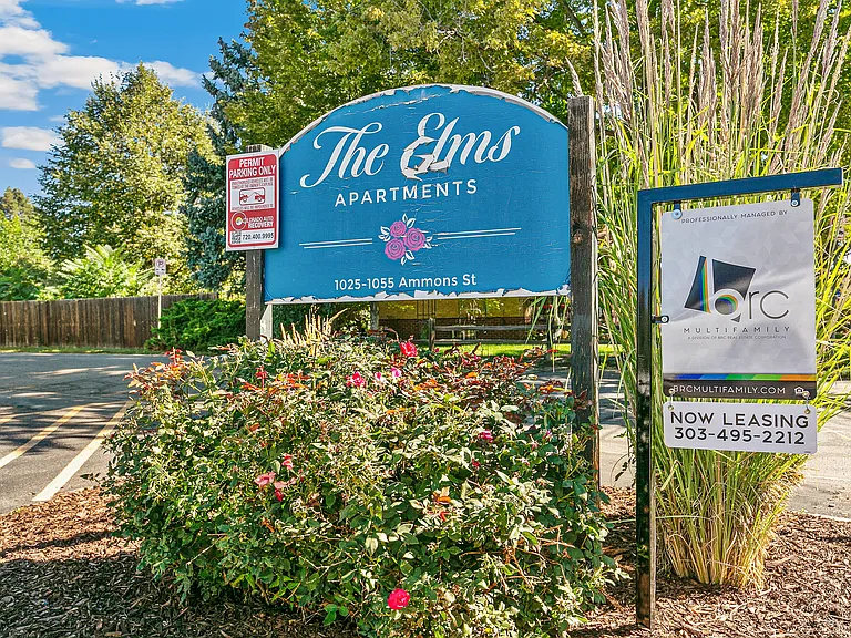 Charming entry sign for The Elms Apartments surrounded by lush greenery and vibrant flowers, located at 1025-1055 Ammons St, offering a welcoming and scenic environment.