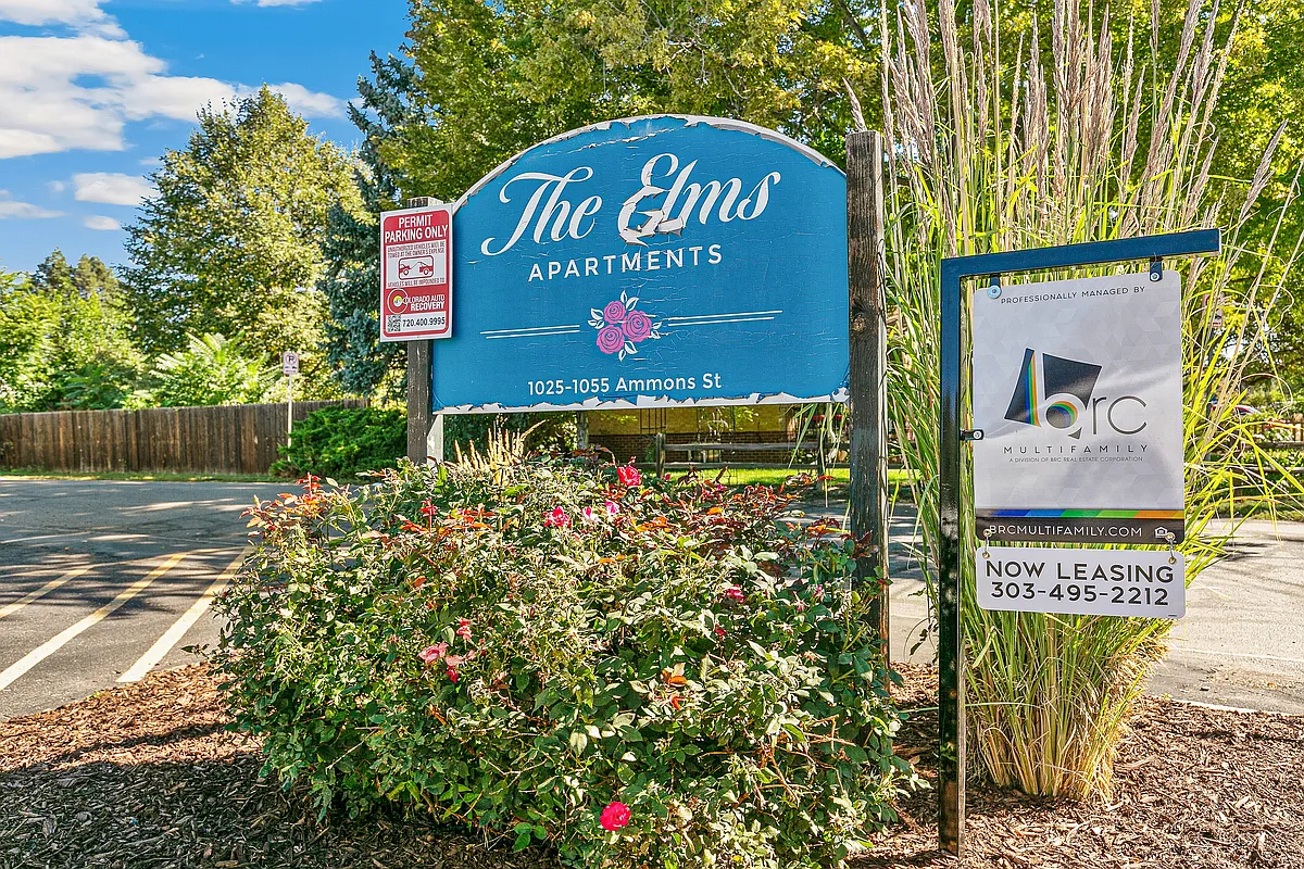 Charming entry sign for The Elms Apartments surrounded by lush greenery and vibrant flowers, located at 1025-1055 Ammons St, offering a welcoming and scenic environment. - 1025 Ammons St