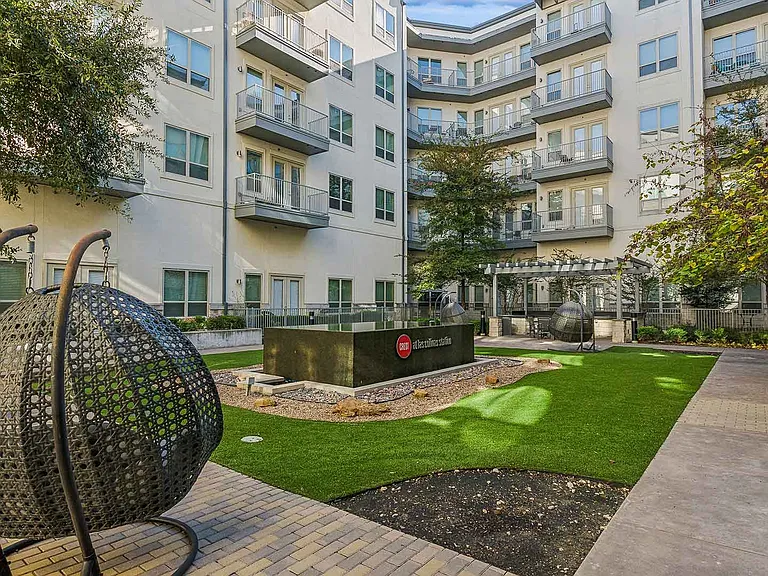 A peaceful courtyard in a residential complex featuring a modern water feature with the Crest Las Colinas logo, surrounded by decorative gravel and vibrant green artificial turf. The space includes un