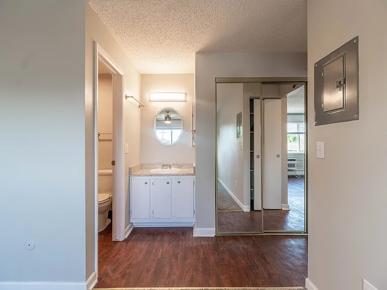 Modern apartment interior featuring bright bathroom with stylish vanity and mirror, mirrored closet doors, and elegant wood flooring, creating a welcoming and airy atmosphere.