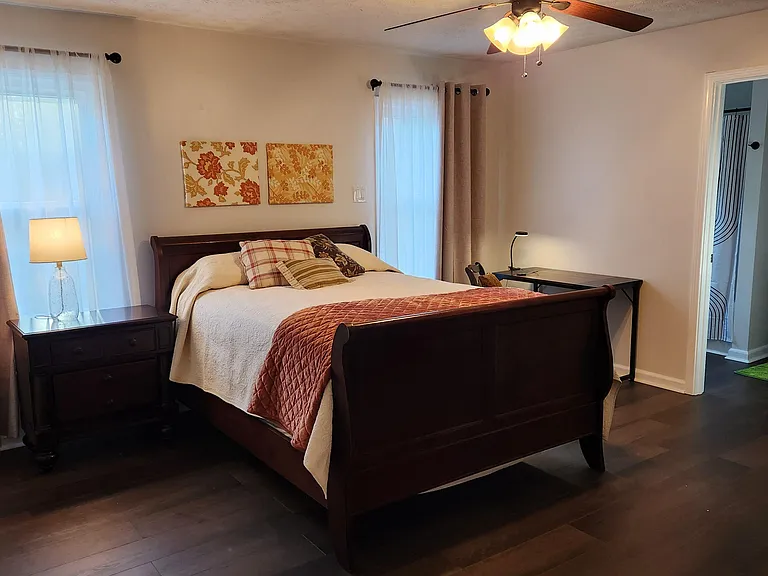 Furnished bedroom with a full bed, nightstand, and dedicated desk area, plus two windows with curtains and ceiling fan.