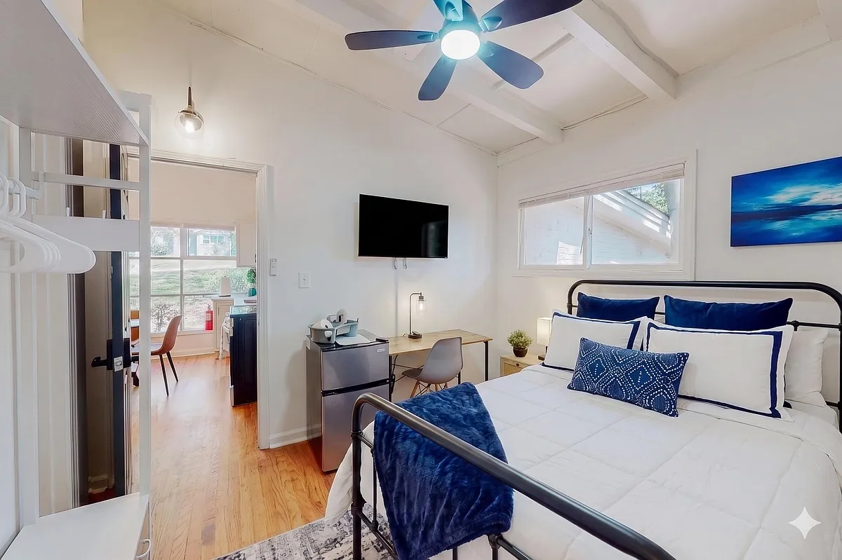 Furnished bedroom with a full bed, ceiling fan, wall-mounted TV, desk, mini fridge, and natural light from large windows. - Monterey Dr