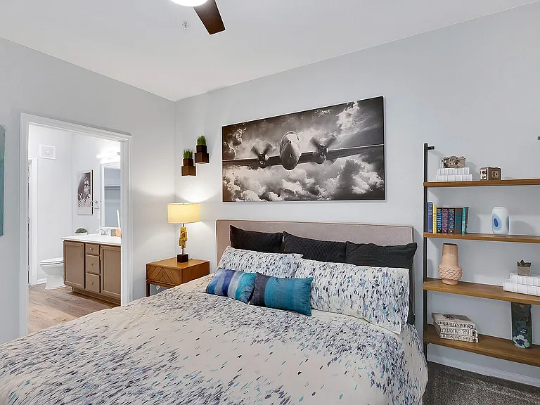 Inviting bedroom with chic decor, featuring a cozy bed with patterned linens, stylish wall art, and a sleek bookshelf, leading to a modern en-suite bathroom.