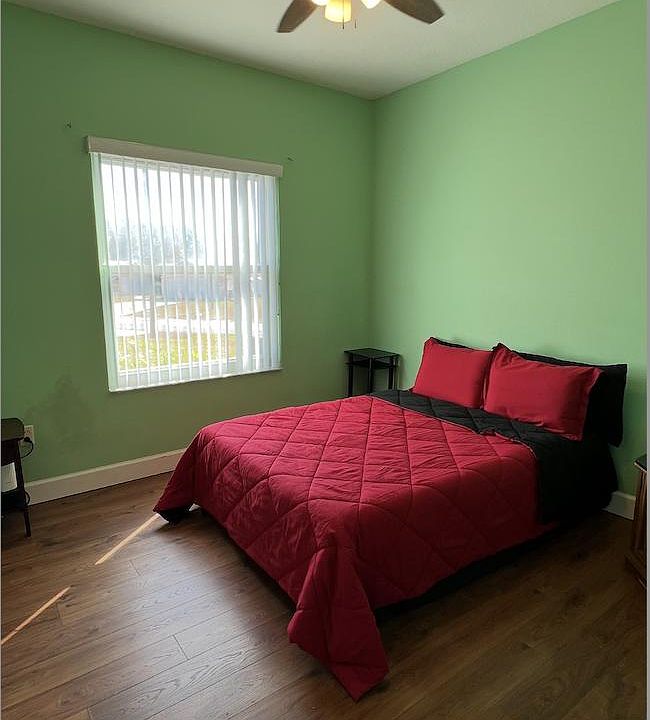Bedroom with wood-style flooring, a ceiling fan, and a large window with vertical blinds for natural light.