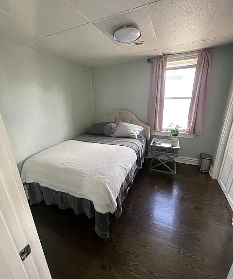 Private bedroom with hardwood floors, a furnished bed, and a window with curtains plus a side table for essentials.