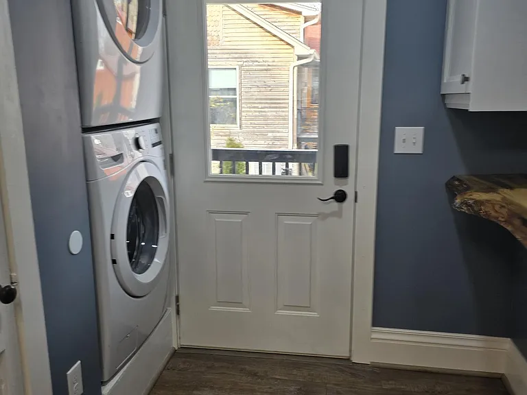 Full sized stacking washer and dryer next to half bath/powder room. View from kitchen. Views of side porch entry with keypad lock. Note live edge shelf on right under cabinet.