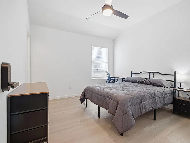 Private bedroom with a full bed, nightstand lamp, and a dedicated desk area, plus a ceiling fan and window blinds.
