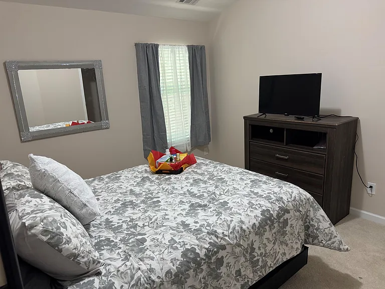 Furnished bedroom with a queen size w/sheets & comforter, Chest, TV, window with curtains for comfortable everyday living.