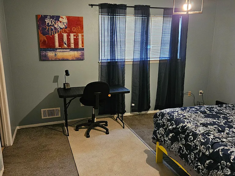 Private bedroom with a full-size bed, dedicated desk workspace, and a window with blinds and curtains for light control.