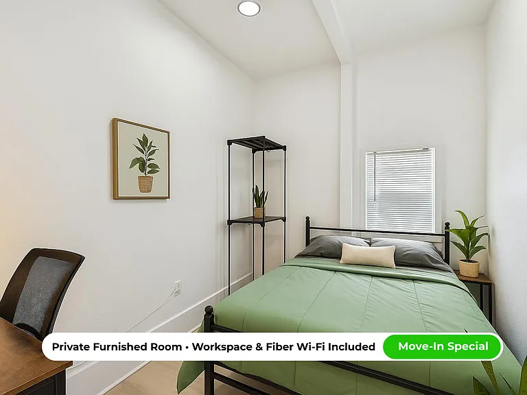 Furnished bedroom with a full-size bed, side table, and dedicated desk area, plus a window with blinds for privacy.