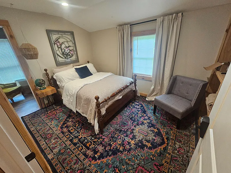 Furnished bedroom with a full bed, nightstand, accent chair, and a window with curtains for natural light and privacy.