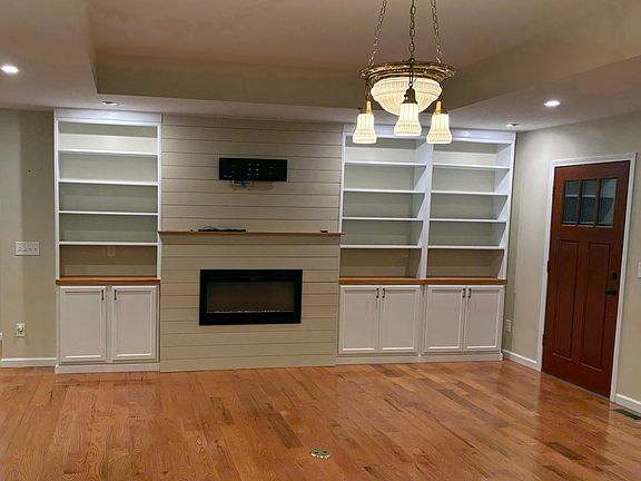 Open living room/kitchen with built in shelving and electric fireplace (with remote), TV mount, tray ceiling, antique light fixture, and hardwood floors.