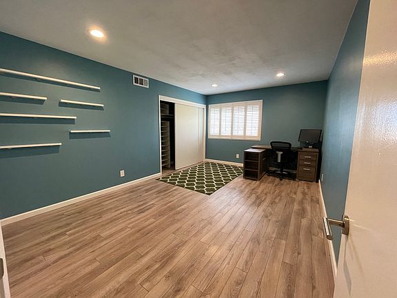 View of room-for-rent from the doorway, with L-desk, closet shelving, and photo mounts on wall