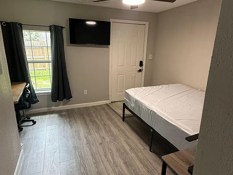 Furnished bedroom with wood-look flooring, ceiling fan, wall-mounted TV, desk workspace, and window with curtains.