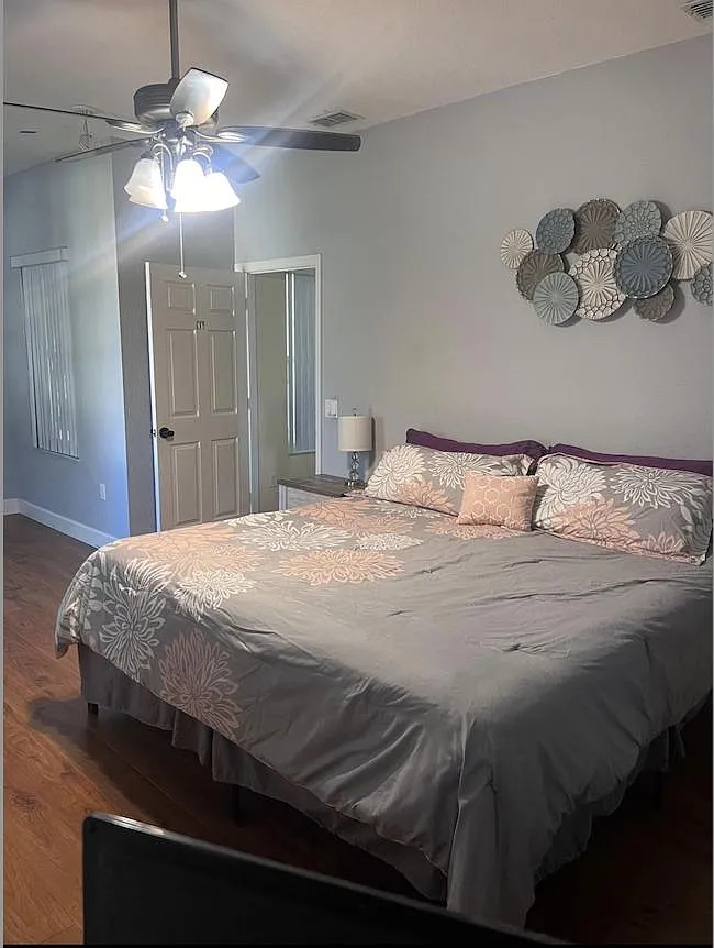Spacious bedroom with a ceiling fan, wood-look floors, and a large window with blinds for privacy and natural light. - Blythe Ave