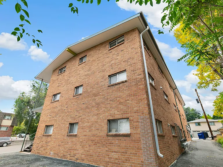 Charming brick apartment building with a vibrant exterior, surrounded by lush trees and a clear blue sky, offering a serene and inviting living environment.