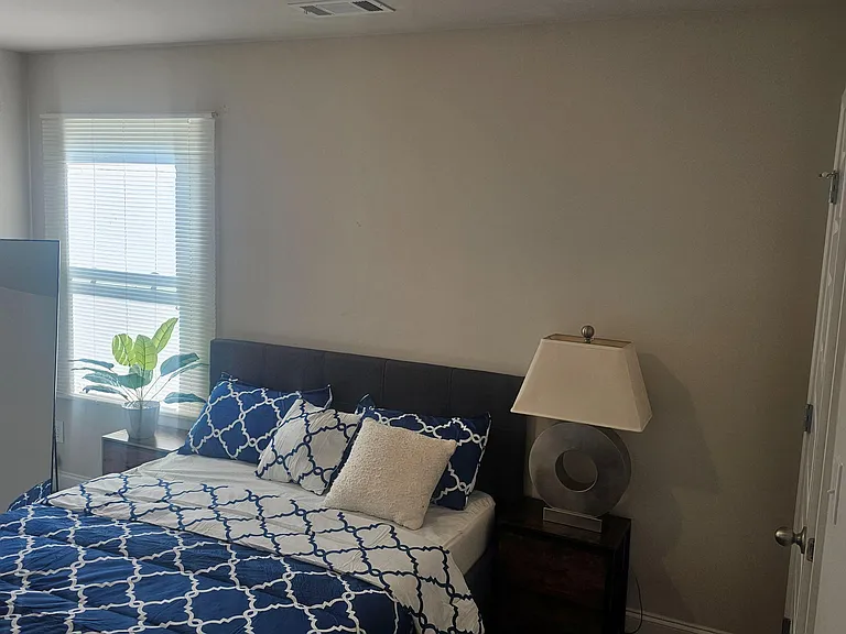 Furnished bedroom with a full/queen bed, nightstand lighting, and two windows with blinds for natural light.