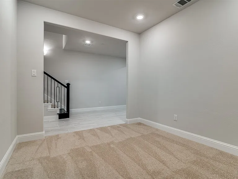 Carpeted room featuring a wide doorway opening to a hall with light-toned flooring, a staircase with black metal railings, and recessed lighting