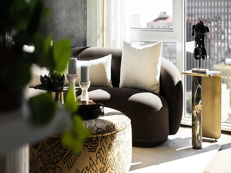 Elegant living room with a modern curved sofa, plush pillows, and a stylish ottoman in front of a bright window, featuring chic decor and contemporary art.