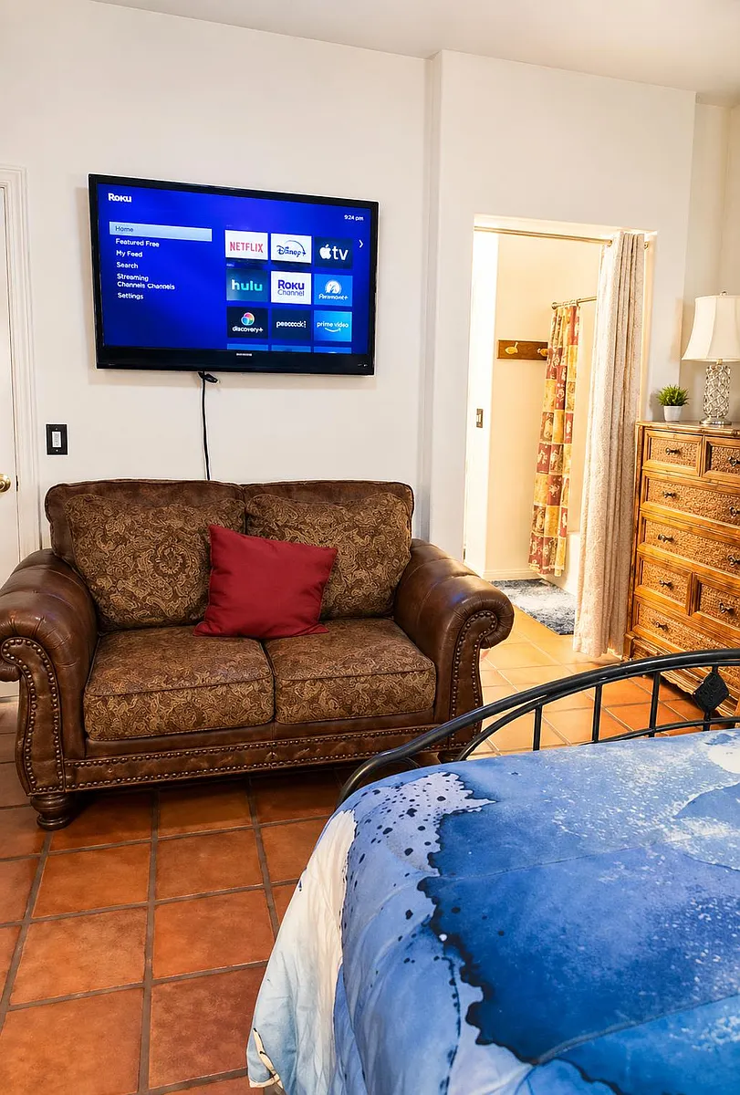 Spacious bedroom suite with a comfortable seating area, wall-mounted TV, and convenient access to the nearby bath area. - N 25th Ave