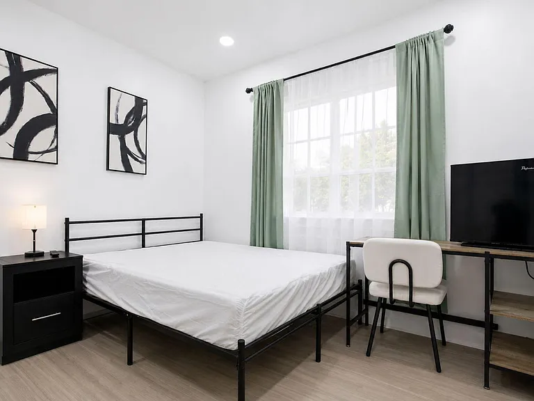 Furnished bedroom with large window, bed and nightstand, plus a desk setup with chair and TV for work or relaxing.