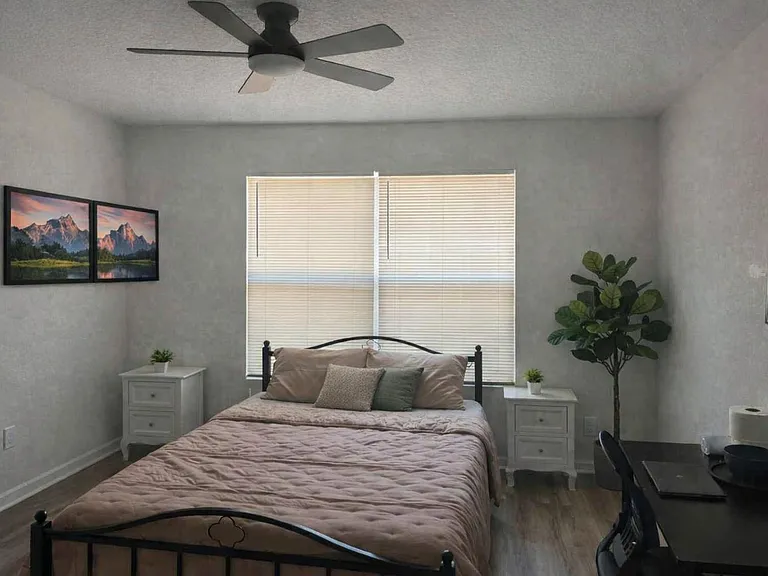 Furnished bedroom with queen bed, dual nightstands, ceiling fan, and a dedicated desk area beside a large window.