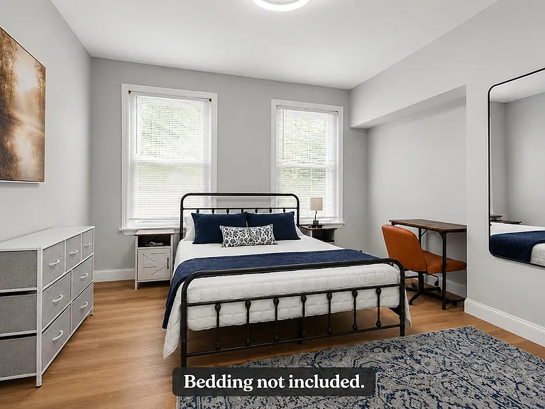 Spacious bedroom with two large windows, a metal bed frame, dresser storage, and a dedicated desk/work area.
