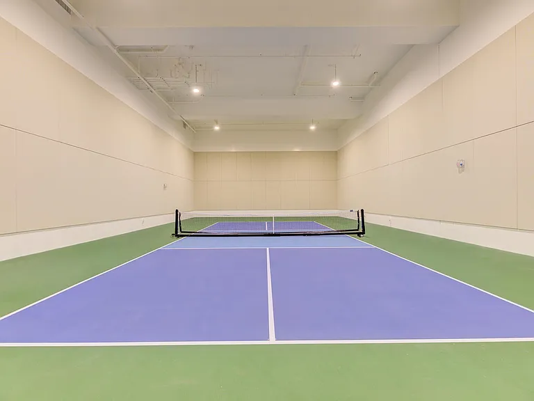 Indoor tennis court with blue flooring and a single net in the center. The walls are light-colored, and the space is brightly lit, creating a clean and spacious environment for playing tennis or pickleball.