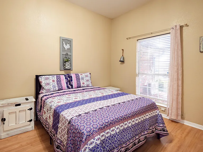 Well-kept bedroom with a comfortable bed, nightstand storage, and a large window with blinds and curtains for privacy.