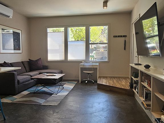 Living room features built-in bookcases, small work desk, sleeper-sofa, large Smart TV, stained concrete flooring with tiled entry. These front windows overlook the courtyard.