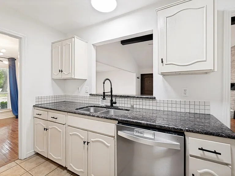 Kitchen featuring light-toned cabinetry, a dark speckled countertop, a stainless steel double sink with a matte black faucet, a white square tile backsplash, and a stainless steel dishwasher